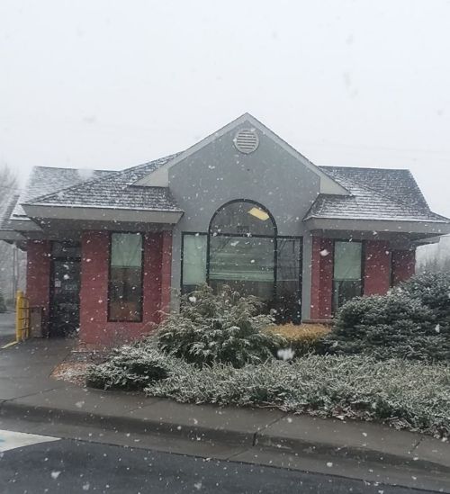 A snow-covered building with a peaked roof and large windows, surrounded by greenery on a winter day.