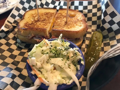A grilled sandwich cut in half, served with coleslaw and a pickle on a checkered paper plate.