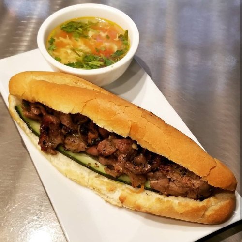 A grilled meat sandwich in a bun with cucumber, served alongside a small bowl of soup with vegetables.