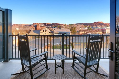 Two rocking chairs and a small table on a balcony overlooking a scenic view of mountains and buildings.