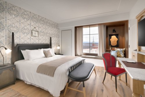 A stylish hotel room featuring a bed, desk, and large window with a scenic view. Elegant decor and warm lighting.