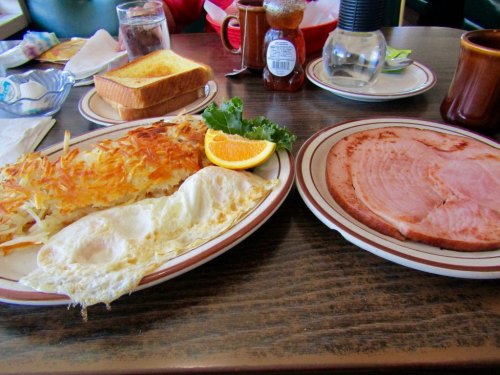 A breakfast plate with eggs, hash browns, ham, toast, and an orange slice on a wooden table.