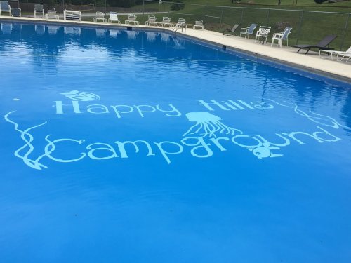 A swimming pool with "Happy Hills Campground" written on the surface, surrounded by lounge chairs and greenery.