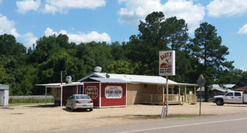 A small BBQ restaurant with a sign, surrounded by trees and a gravel parking lot, under a blue sky with clouds.