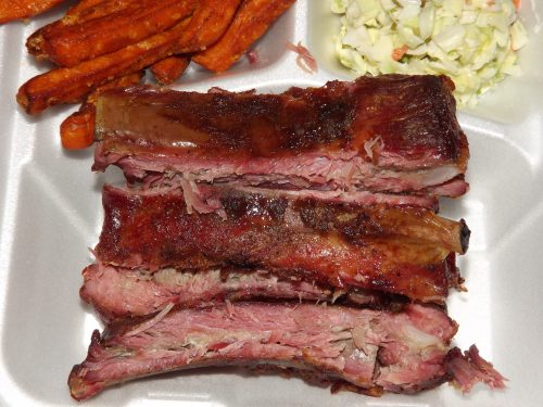 Close-up of tender, smoked ribs with a side of sweet potato fries and coleslaw in a takeout container.