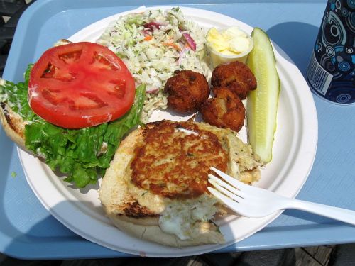 A plate with a sandwich topped with lettuce and tomato, coleslaw, a pickle, hushpuppies, and a dollop of butter.