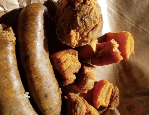 A close-up of a plate featuring sausages, a round meatball, and chunks of sweet potato on brown paper.