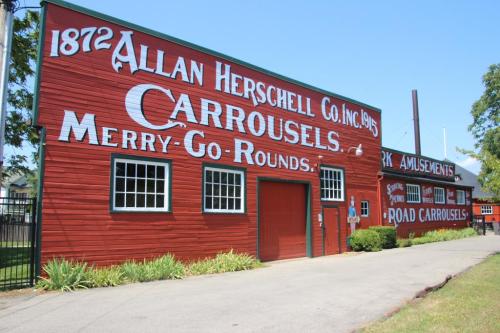 Red wooden building with large white text reading "Allan Herschell Co. Inc. 1872 Carrousels Merry-Go-Rounds."