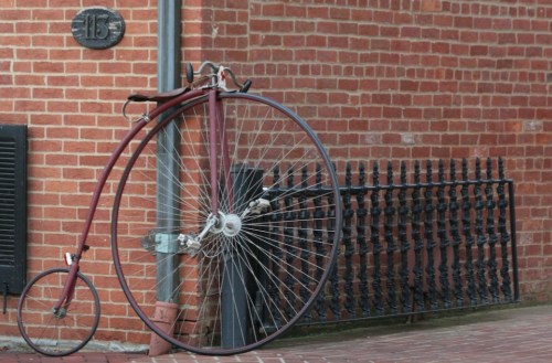 A vintage penny-farthing bicycle leans against a brick wall, with a decorative iron fence in the background.