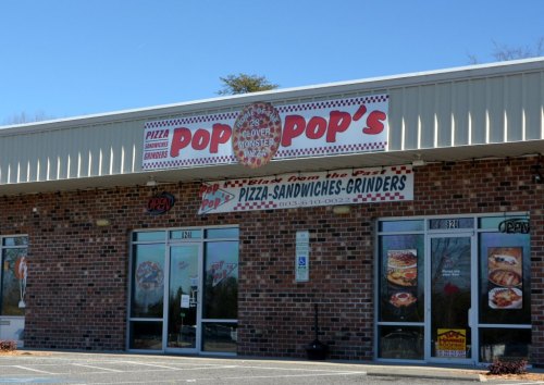 Exterior of Pop's Pizza, featuring a brick facade and signs for pizza, sandwiches, and grinders. Clear blue sky above.