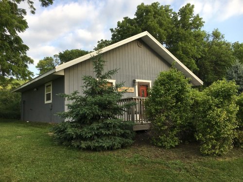 A gray house with a porch surrounded by green trees and shrubs under a partly cloudy sky.