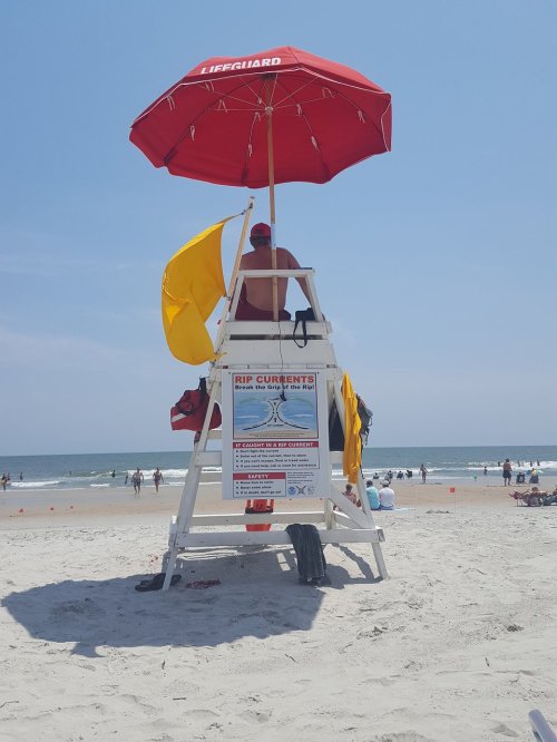 A lifeguard sits on a tall chair under a red umbrella, watching over a busy beach with warning flags and a sign.
