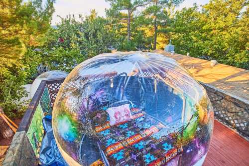 A transparent bubble structure on a deck, surrounded by trees, featuring colorful cushions inside.