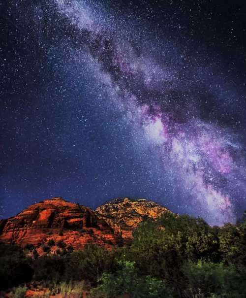 A starry night sky filled with the Milky Way over red rock formations and lush greenery.