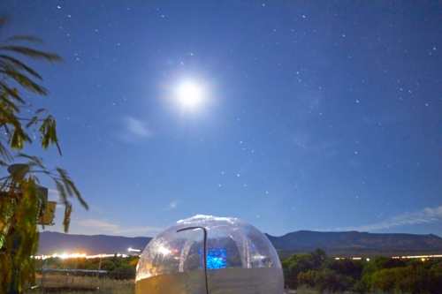 A transparent dome under a bright moon and starry sky, with mountains in the background.