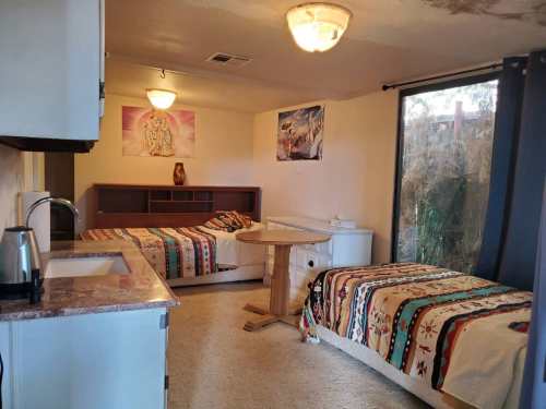 A cozy room with two beds, a round table, kitchenette, and large window with natural light. Decorated with colorful blankets.
