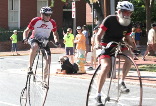 Two cyclists ride on a street; one on a modern bike and the other on a vintage penny-farthing, with spectators in the background.