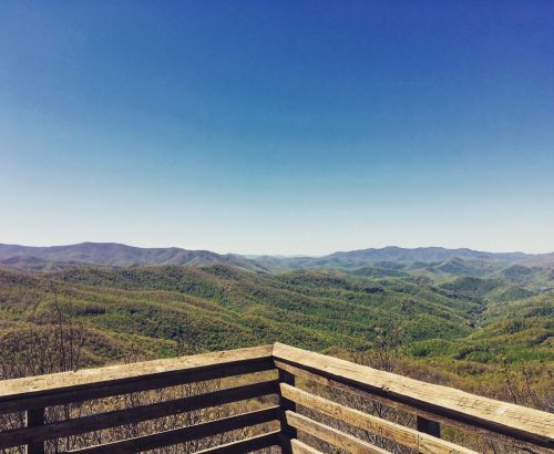 A panoramic view of lush green mountains under a clear blue sky, seen from a wooden overlook.