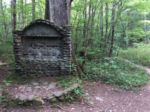A stone monument covered in moss stands in a lush green forest, with a dirt path winding nearby.