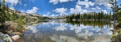 A serene lake surrounded by mountains and trees, reflecting clouds and blue sky in calm waters.
