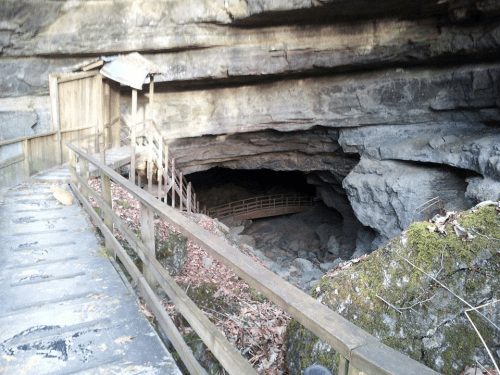 A wooden path leads to a cave entrance, surrounded by rocky cliffs and scattered leaves.