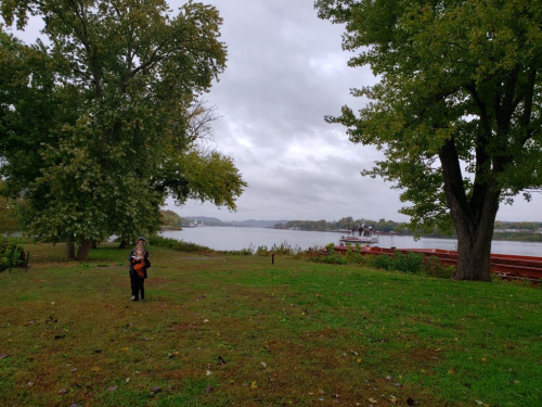 A person stands on a grassy area by a river, surrounded by trees under a cloudy sky. A boat is visible in the distance.