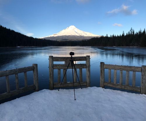 A snowy landscape with a camera on a tripod facing a frozen lake and a snow-capped mountain in the background.