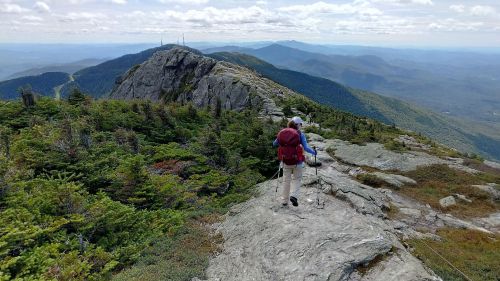 A hiker walks along a rocky trail with lush greenery and distant mountains under a cloudy sky.