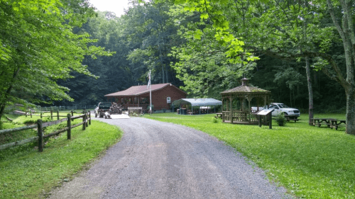 A gravel road leads to a rustic cabin surrounded by trees, with a gazebo and parked vehicles nearby.