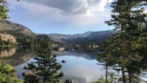 A serene lake surrounded by mountains and trees, reflecting the sky and clouds in calm waters.