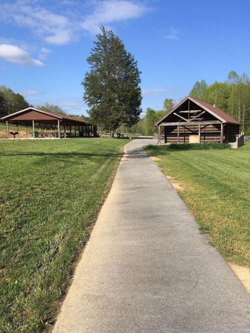 A paved path leads through a grassy area, flanked by two wooden structures under a blue sky with scattered clouds.