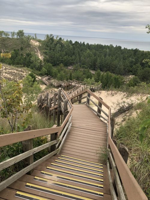 Wooden boardwalk winding down through sandy dunes, leading to a view of the lake and surrounding greenery.