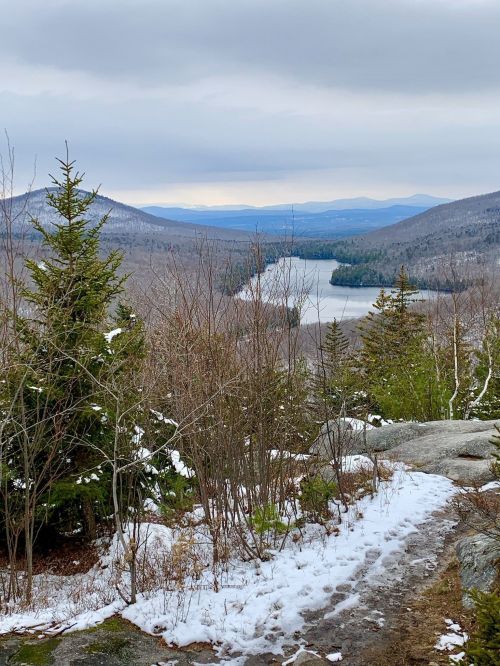 A scenic view of a lake surrounded by mountains and trees, with patches of snow on the ground under a cloudy sky.
