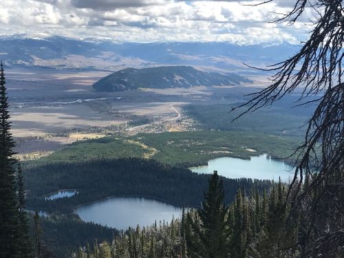 A panoramic view of a valley with lakes, forests, and distant mountains under a cloudy sky.