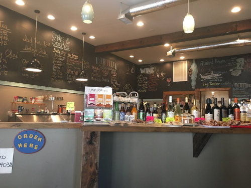 A cozy bar with a wooden counter, displaying various wines and drinks, and a chalkboard menu in the background.