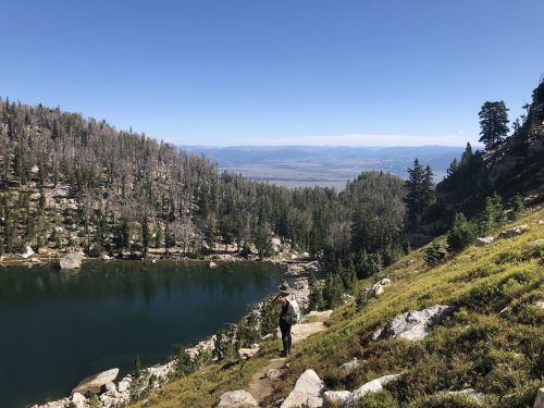 A hiker walks along a rocky path beside a serene lake, surrounded by trees and mountains under a clear blue sky.