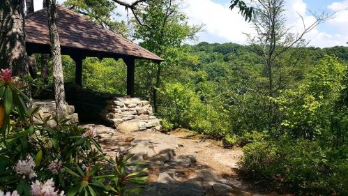 A stone gazebo surrounded by lush greenery and trees, overlooking a scenic landscape under a blue sky.