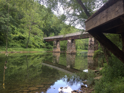 A rustic bridge spans a calm river, surrounded by lush greenery and reflected in the water below.