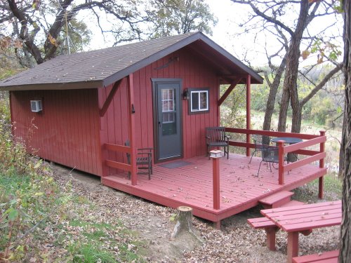 A small red cabin with a porch, surrounded by trees and autumn leaves, set on a hillside.