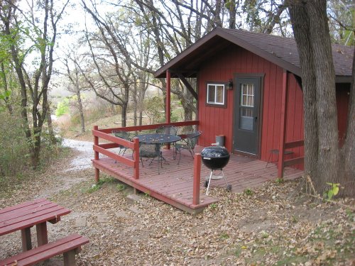 A small red cabin with a porch, surrounded by trees and a picnic table, near a gravel path in a wooded area.