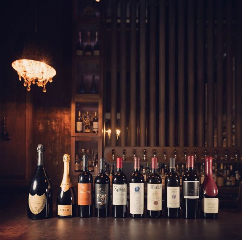 A selection of wine bottles displayed on a bar, with a chandelier and wooden backdrop in a dimly lit setting.