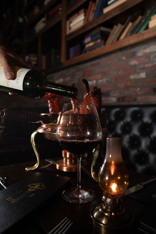 A hand pours red wine into a glass at a dimly lit table, with a candle and books in the background.