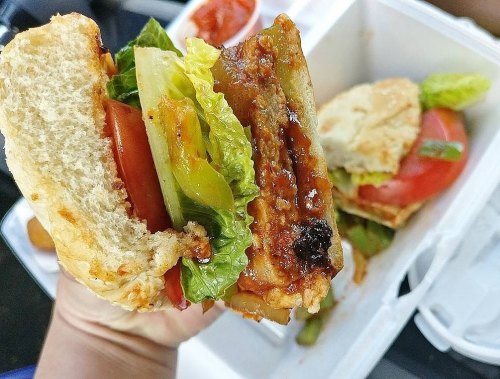 A close-up of a hand holding a sandwich with lettuce, tomato, and a grilled patty, with a side of sauce in a takeout box.