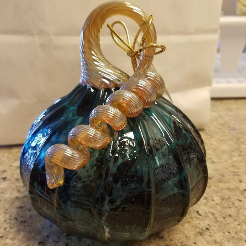 A decorative glass pumpkin with a twisted golden stem, featuring a dark teal and black marbled surface.