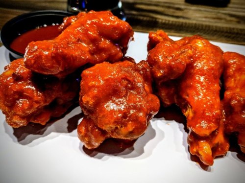 A close-up of spicy chicken wings coated in red sauce, served with a small cup of dipping sauce on the side.