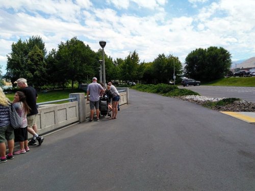A group of people stands near a railing in a park, with trees and a parking lot visible in the background.