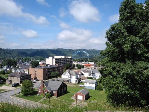 A scenic view of a town with houses, a bridge in the background, and green hills under a partly cloudy sky.