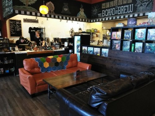 Cozy café interior with a brown couch, wooden tables, and shelves of board games; staff at the counter in the background.