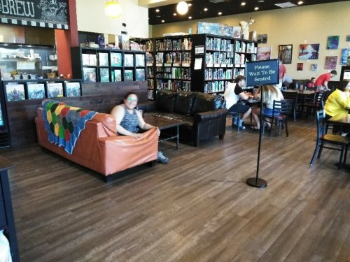 A cozy café interior with a couch, colorful blanket, bookshelves, and patrons seated at tables. A sign requests to wait to be seated.