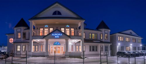 A large, well-lit inn with a sign reading "Aurora Inn," surrounded by snow and a fence, at dusk.
